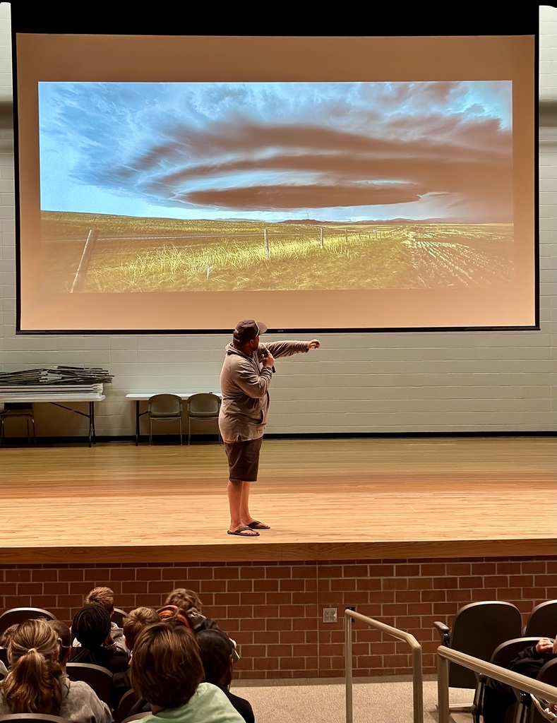 Chris Chittick stands in front of a projected image of a tornado while speaking to sixth-grade students seated in the auditorium.