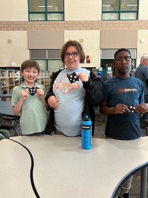 Three students stand together in the library holding black bat-shaped origami bookmarks and smiling for the camera.