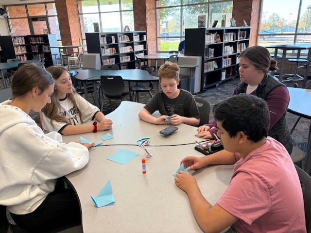 A small group of students sits at a table folding blue paper into origami shapes during the Best Buddies craft activity.