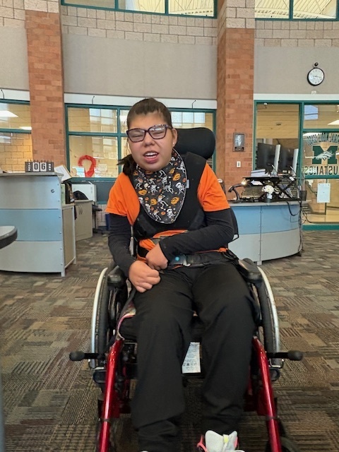 A student wearing an orange and black Halloween shirt smiles while seated in a wheelchair, showing off his completed craft.