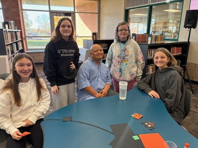 Five students from the Best Buddies program sit together at a table in the library, smiling while working on Halloween crafts.