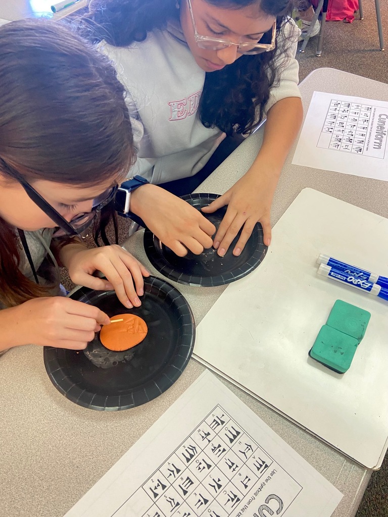 A student presses a stylus into a clay tablet to write in cuneiform while sitting at a classroom table. Other clay tablets and tools are visible on the desk.