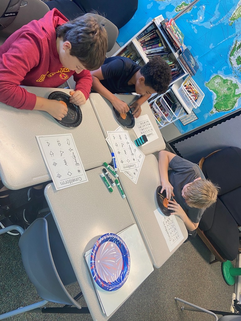 Two students sit side by side, carving wedge-shaped symbols into clay tablets as part of a classroom activity about Mesopotamian writing.
