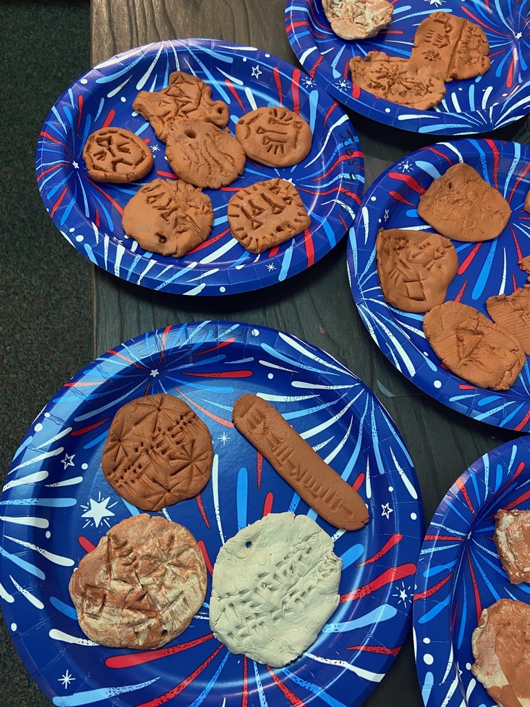 Several completed clay tablets with cuneiform writing rest on a classroom table beside sculpting tools and reference sheets.
