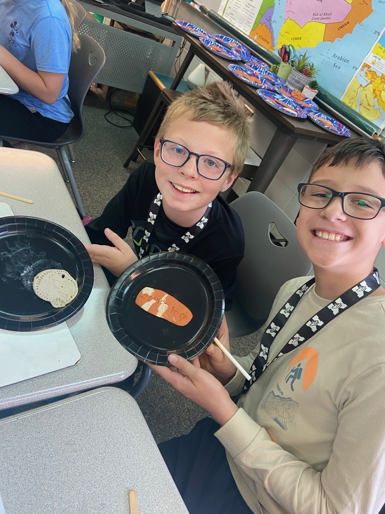 Two students sit side by side, carving wedge-shaped symbols into clay tablets as part of a classroom activity about Mesopotamian writing.