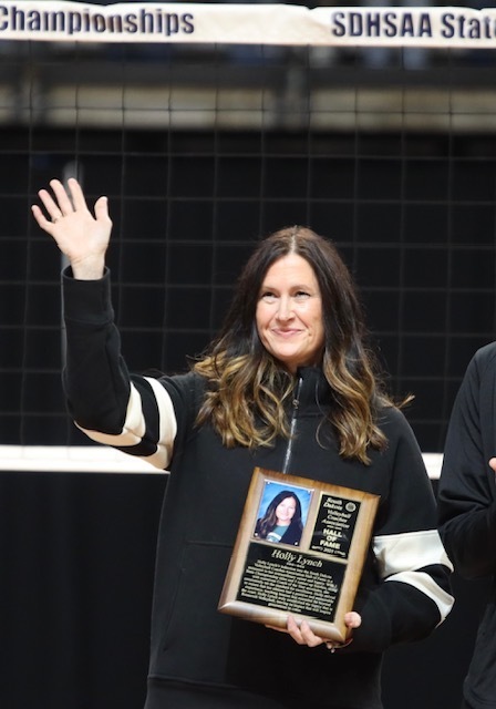 Holly Lynch stands on a volleyball court, smiling and waving with one hand while holding a Hall of Fame plaque in the other. She is wearing a black jacket and standing in front of a net during an induction ceremony.