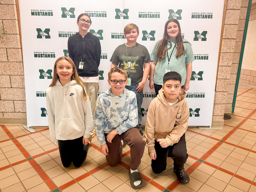 A group of smiling sixth graders stand and kneel in front of the Mustangs backdrop to celebrate their selection as students of the month.
