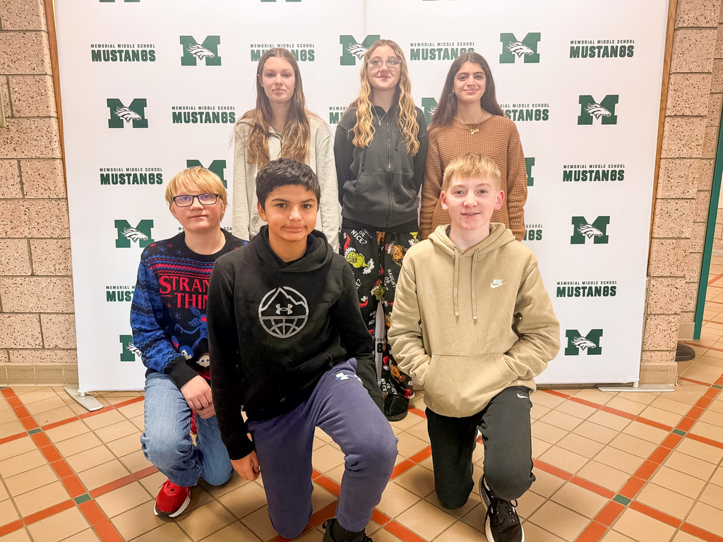 A group of smiling seventh graders stand and kneel in front of the Mustangs backdrop to celebrate their selection as students of the month.