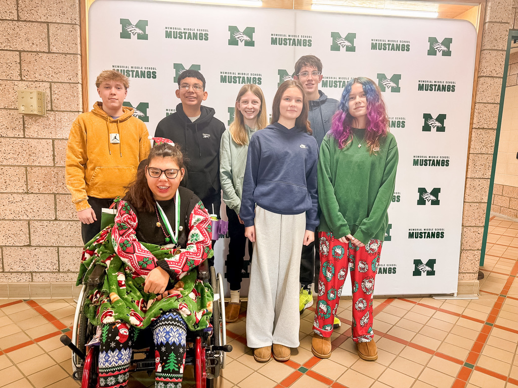 A group of smiling eighth graders stand in front of the Mustangs backdrop and a RISE student is seated in her wheelchair to celebrate their selection as students of the month.