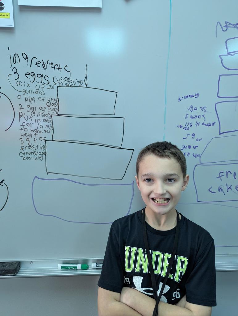 Student smiles with arms crossed in front of a whiteboard showing a drawn tiered cake and handwritten “friendship recipe” ingredients.