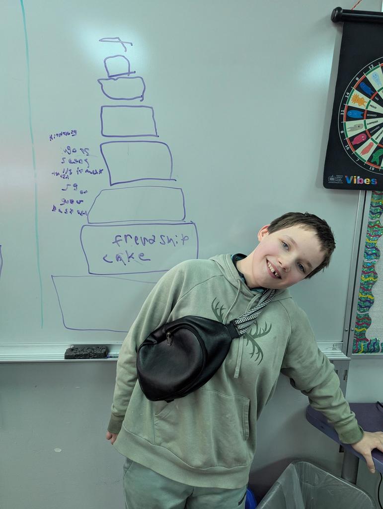 Student smiles while leaning sideways in front of a whiteboard drawing of a multi-layer “friendship cake” with the words “friendship cake” written on the bottom layer.