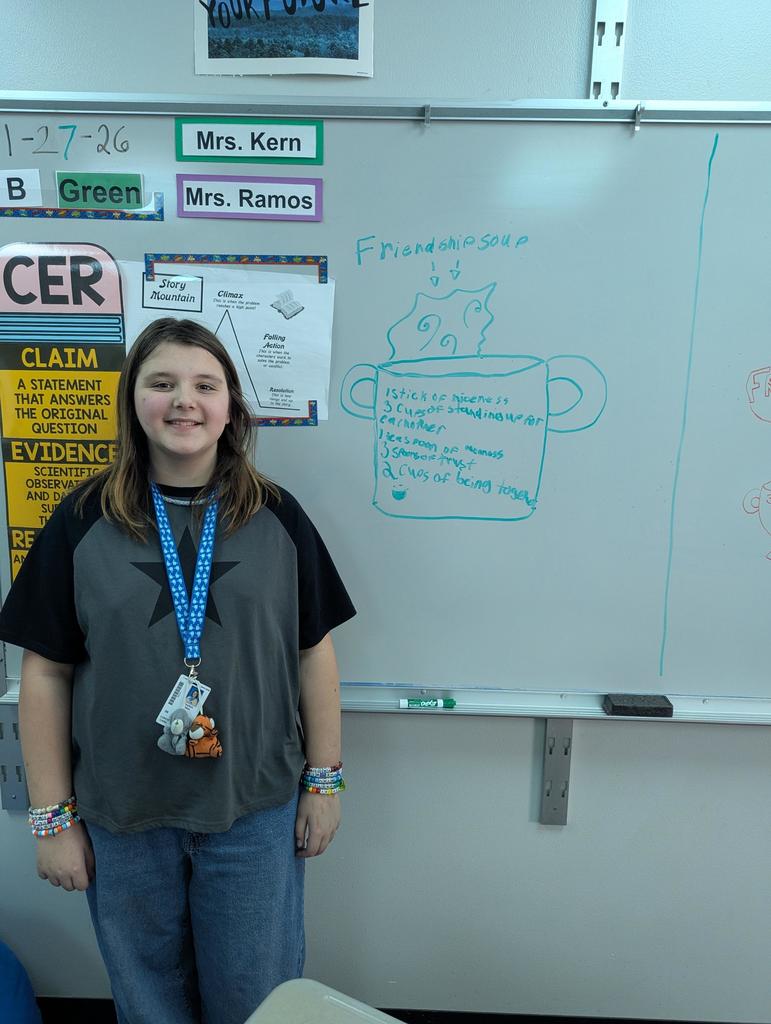 Student stands smiling in front of a whiteboard drawing labeled “Friendship soup,” featuring a pot illustration and a handwritten list of friendship ingredients.