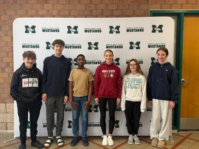 Six 8th grade Students of the Month stand in a line in front of a Memorial Middle School Mustangs step-and-repeat backdrop in a school hallway.