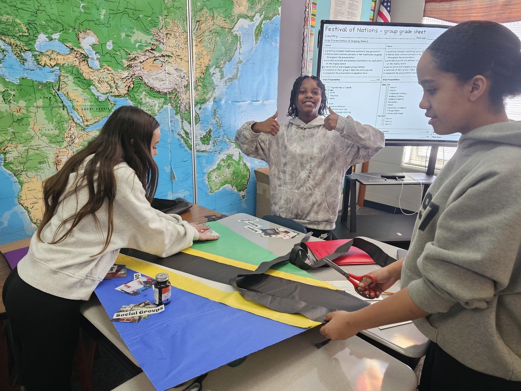 Three students work around a table assembling a large flag display with colored paper. One student uses scissors while another smooths paper on the table. A world map and a projected “Festival of Nations” grading sheet are visible behind them.
