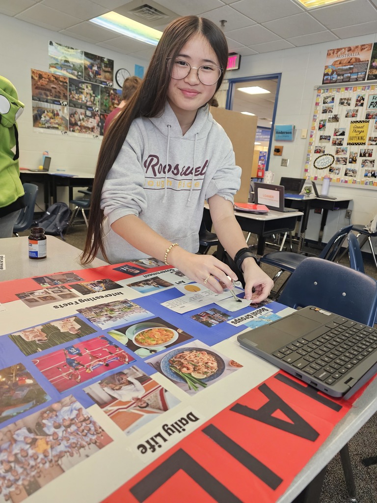 Student in a classroom works at a table assembling a tri-fold presentation board with photos and headings for a country project. A laptop sits nearby, and classroom posters and student work are visible in the background.