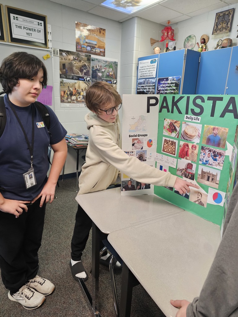 Two students stand beside a tri-fold presentation board labeled “PAKISTAN.” One student points to a section on the board while the other looks on. The classroom walls display photos and geography-related posters.