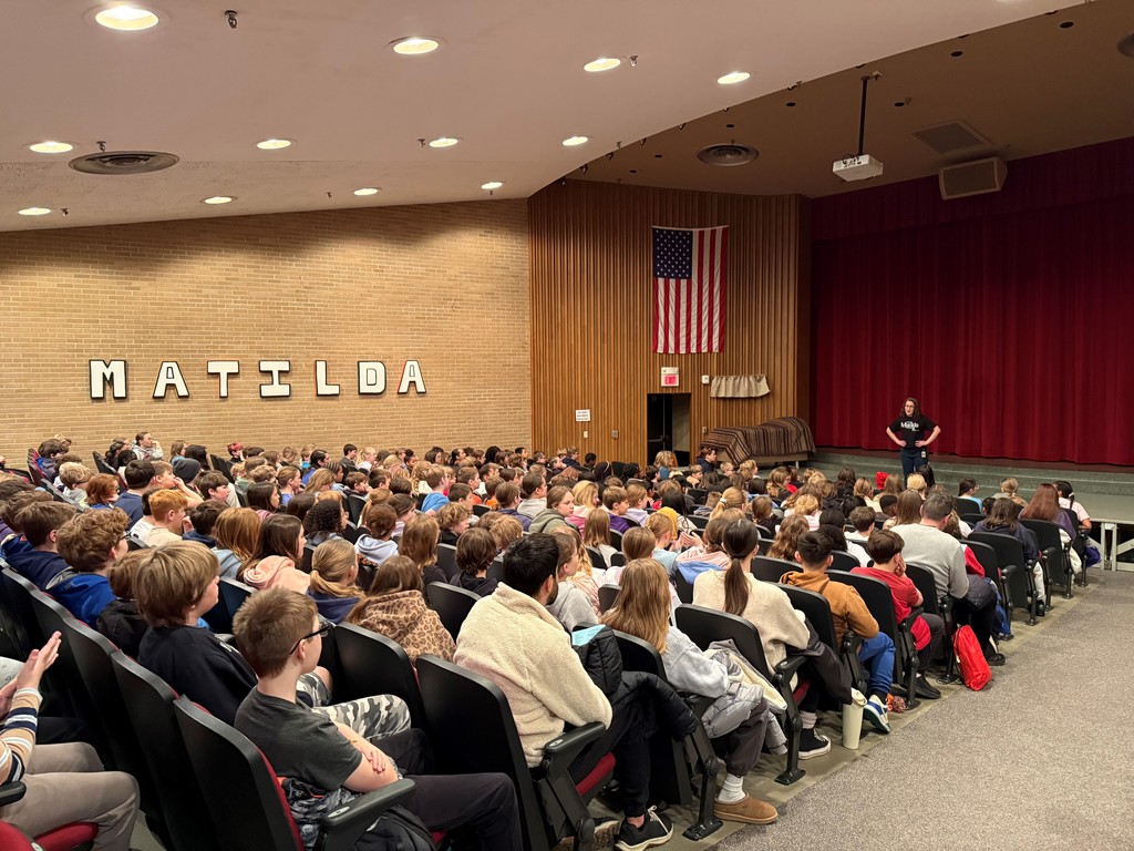 A group of elementary students seated in an auditorium watching a school performance of Matilda the Musical. The stage is set for the production, and performers are visible under bright stage lights. The audience of 5th graders appears engaged and excited, watching attentively and enjoying the show.