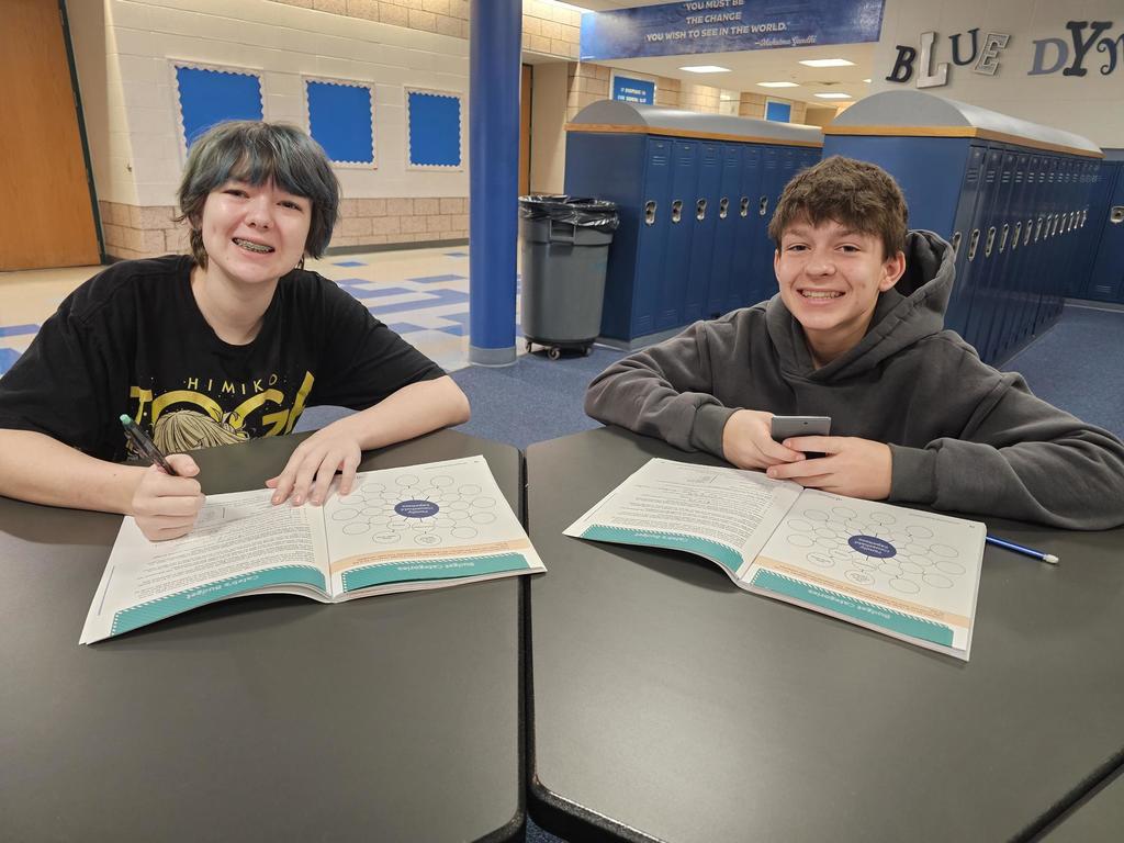 Two students sit at separate tables in a school hallway with budgeting worksheets open. Lockers line the hallway behind them.