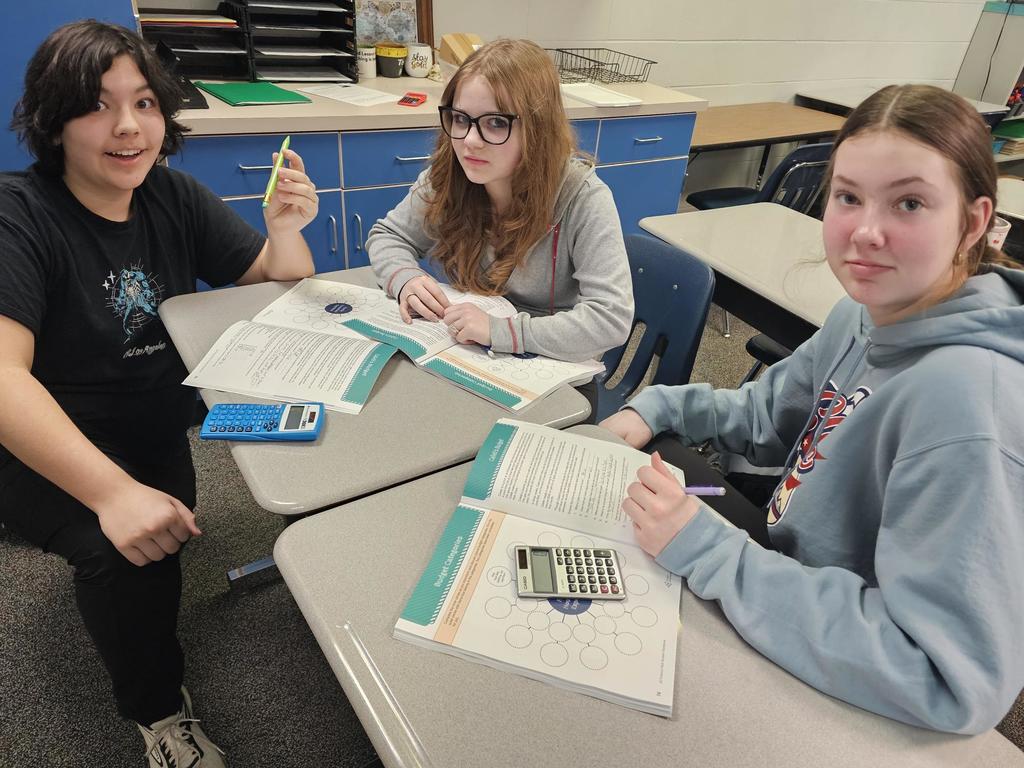 Three students sit at grouped desks with budgeting worksheets open. One student holds a pencil while two calculators sit on the desks.