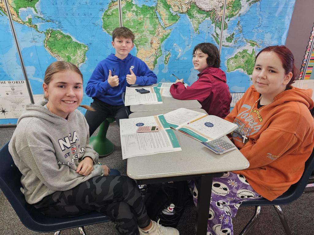 Four students sit around a table with budgeting worksheets open. A large world map covers the wall behind them, and calculators are on the table.