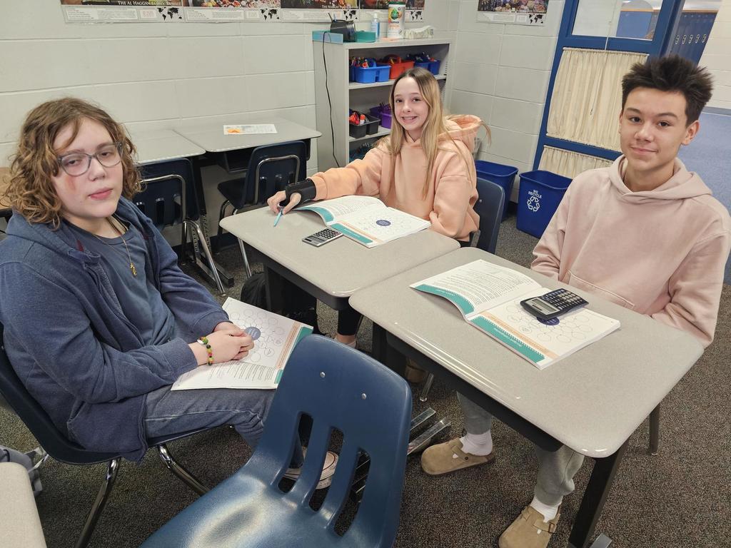 Three students sit at separate desks facing the camera with JA budgeting worksheets open and a calculator on one desk. Classroom shelves and supplies are in the background.