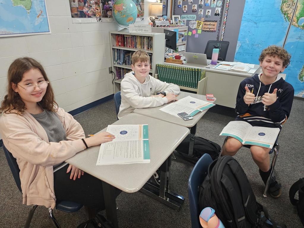 Three students sit at desks with budgeting worksheets open and calculators nearby. A classroom globe, bookshelves, and world map are visible behind them.