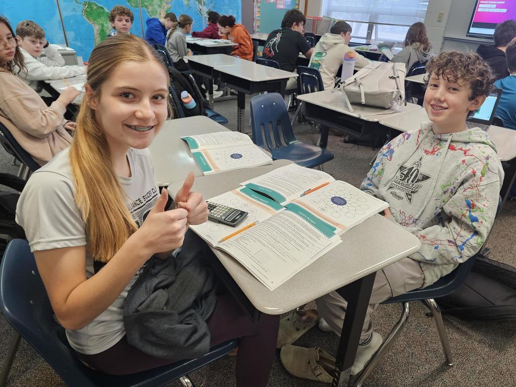 Two students sit at a table with budgeting worksheets open. One student gives a thumbs up while the class works at desks in the background.
