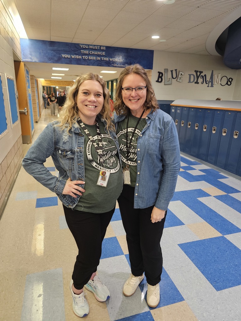 Two staff members smile in a Memorial Middle School hallway lined with blue lockers. Both wear Memorial green “Mustangs” shirts and denim jackets, standing under a hallway quote and a “Blue Dynamos” sign.