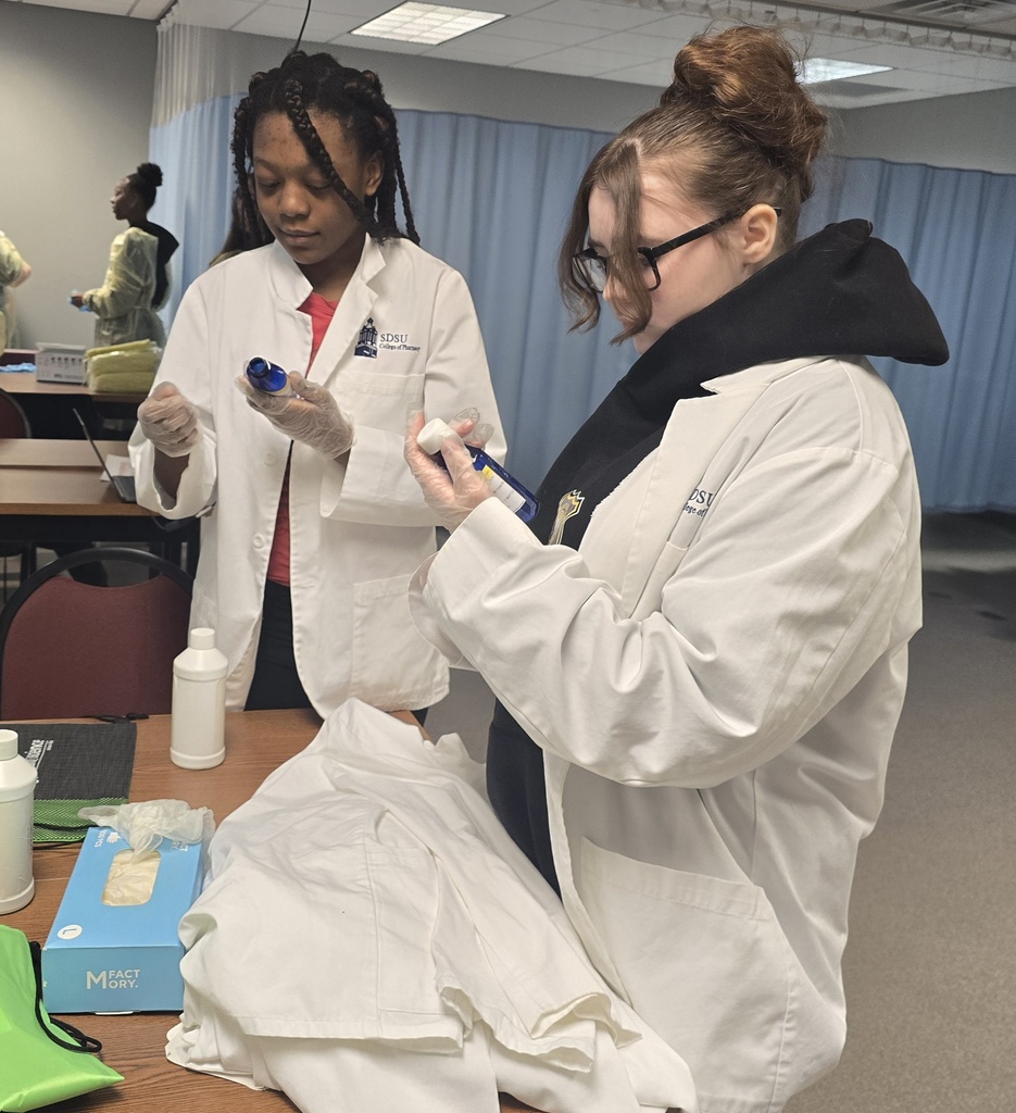 Two students in white lab coats looking at blue glass bottles.