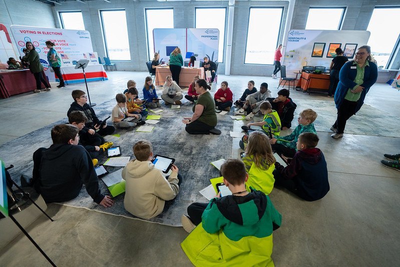 Group of middle school students sit in a circle with their attention on a woman in the middle of the circle talking