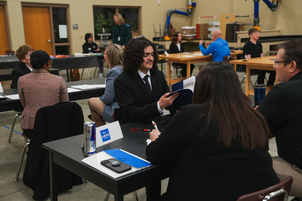 Academy of Finance student in a suit and tie opens a folder with colorful graphs to show two LEVO Credit Union representatives during a mock interview at the CTE Academy.