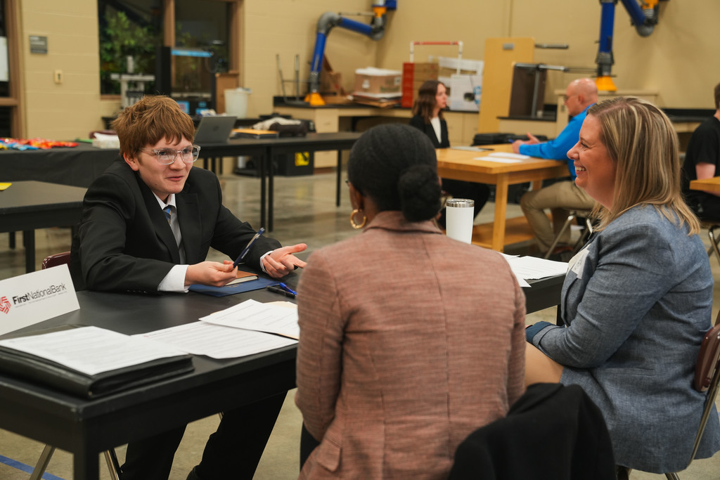 Academy of Finance student in a suit and tie sits at a table gesturing confidently at two First National Bank representatives during a mock interview at the CTE Academy.