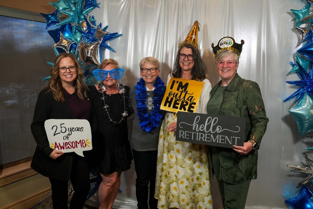 Five Memorial Middle School staff members pose together at a retirement celebration in front of a white curtain backdrop with blue and silver star balloons. From left to right: Patty Hansen holds a sign reading "25 Years of Awesome"; Julie Carlson wears oversized blue novelty glasses and a star necklace; Deb Karlson wears a blue lei and a gold party hat; Sue Zueger holds a yellow sign reading "I'm Outta Here"; and Julie Ortman wears a "The Legend Has Retired" crown and holds a sign reading "Hello Retirement."