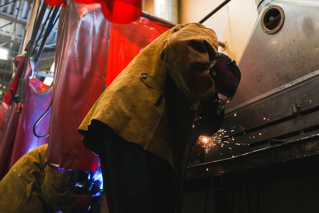 A student in a heavy suede jacket welds as sparks emit from the torch