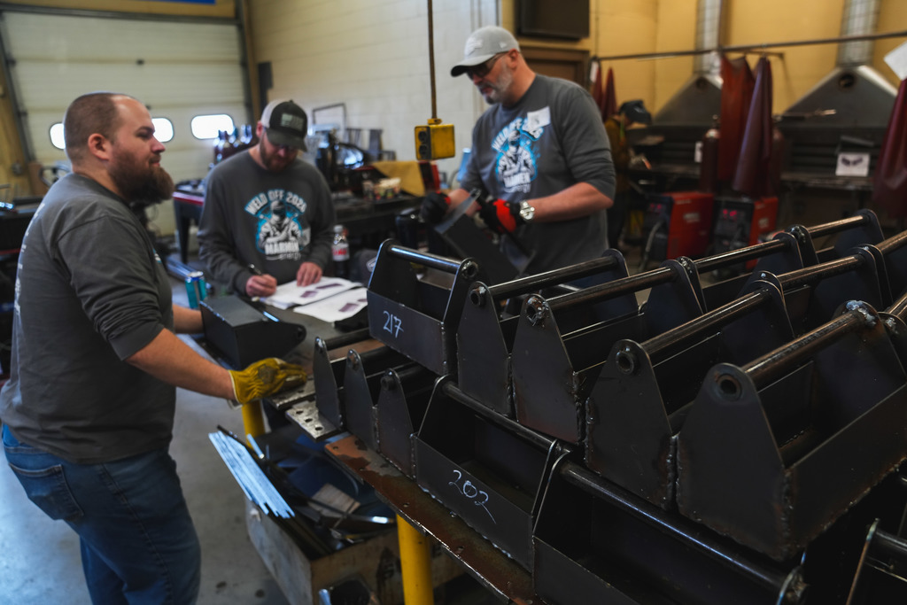 A group of welded tool boxes sits stacked in the foreground while a group of 3 men judge more welded tool boxes in the background