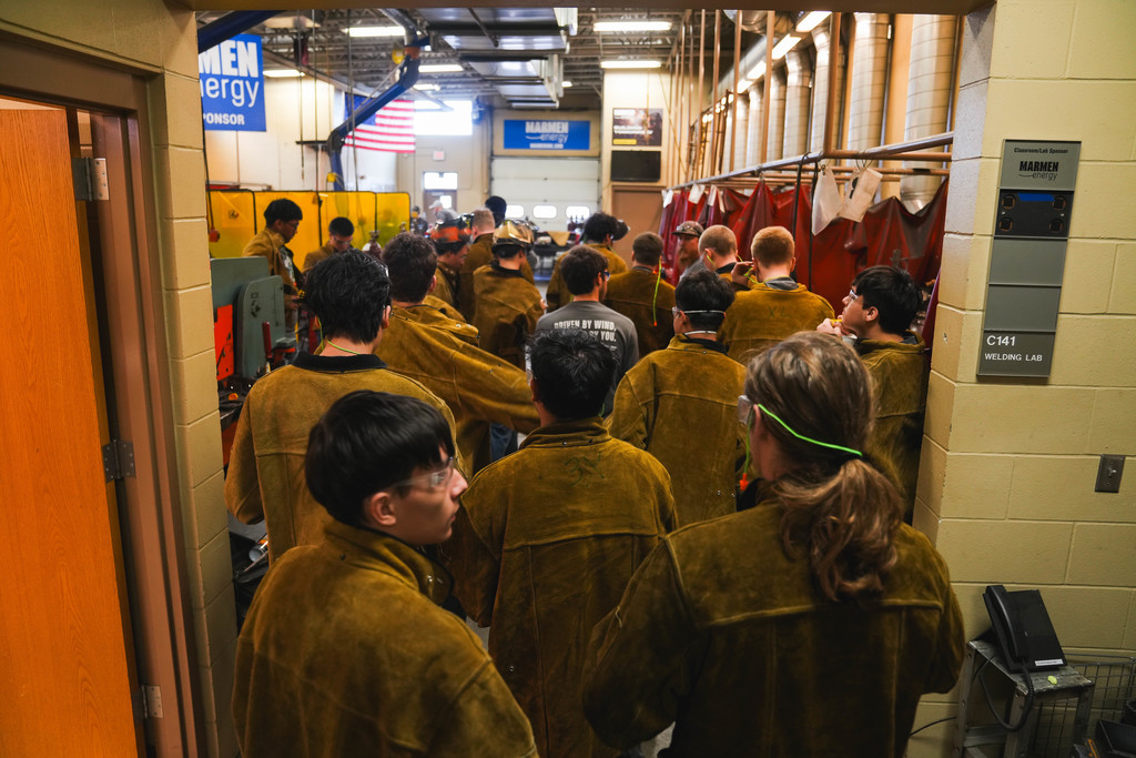 A group of students walks into a welding lab all wearing heavy suede leather jackets. the backs of their heads are visible 