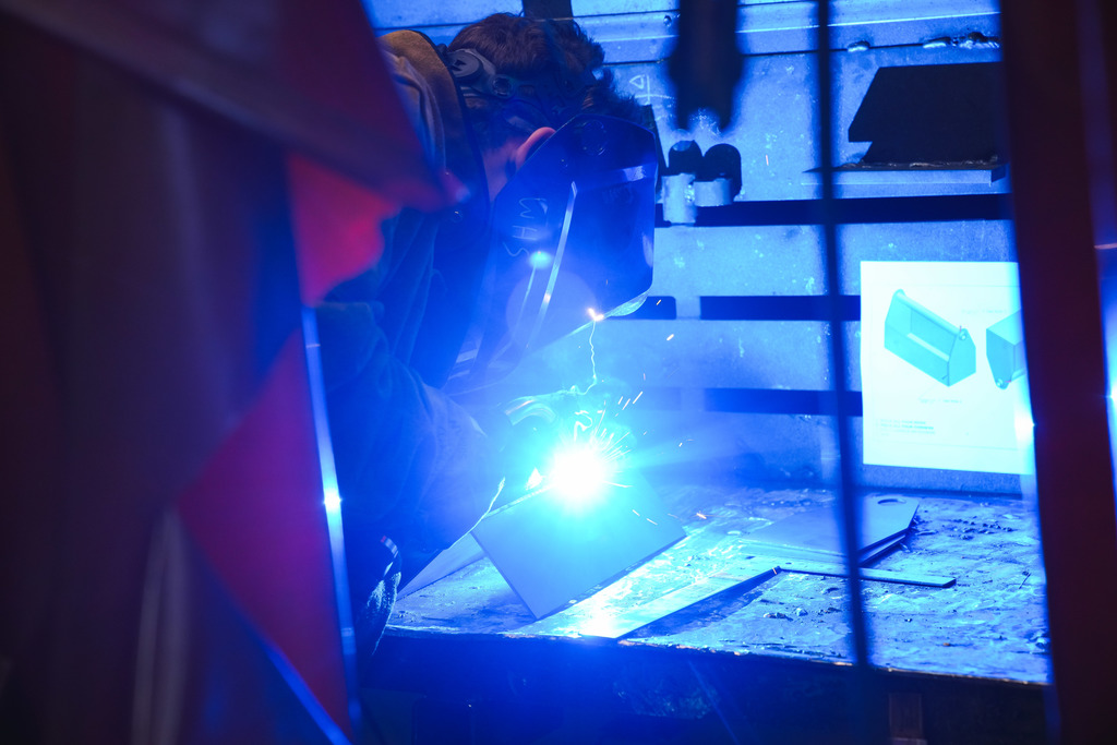 A student with a welding mask welds 2 pieces of metal together. A bright blue light emits from the weld. 