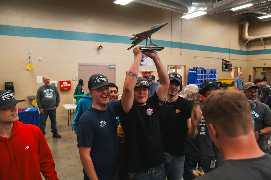 A group of students in hats hold up a trophy of an airplane high above their heads 