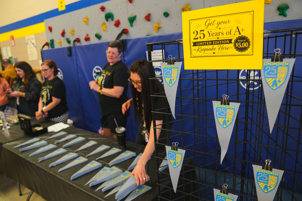 A sign says "Get your 25 years of A+ limited edition keepsake here" in the forground with teachers selling triangle pendants in the background