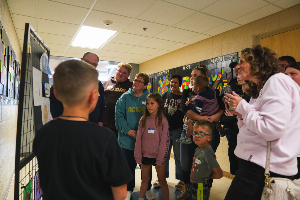 We see the back of a young boys head while a large group of family members listen to him talk and explain his art. We can see their faces and they are happy to listen. 