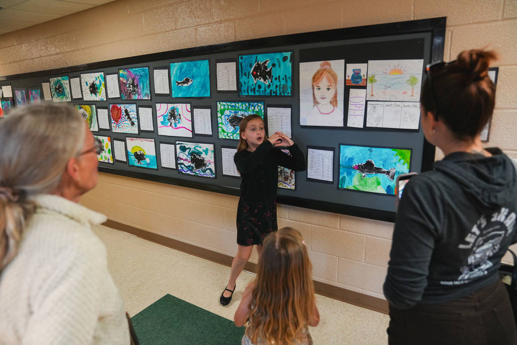 A young girl makes a sign with her hand to explain art that hangs behind her to a group of adult woman with a small child with long hair 