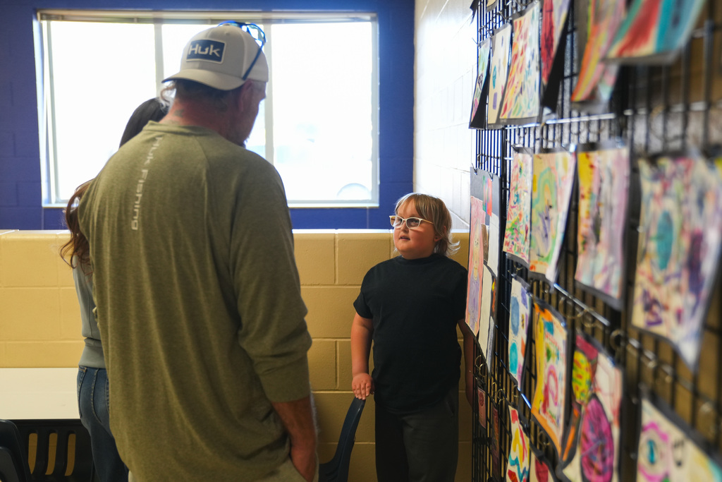 A young boy with glasses talks to two adults in front of a wall of kids art