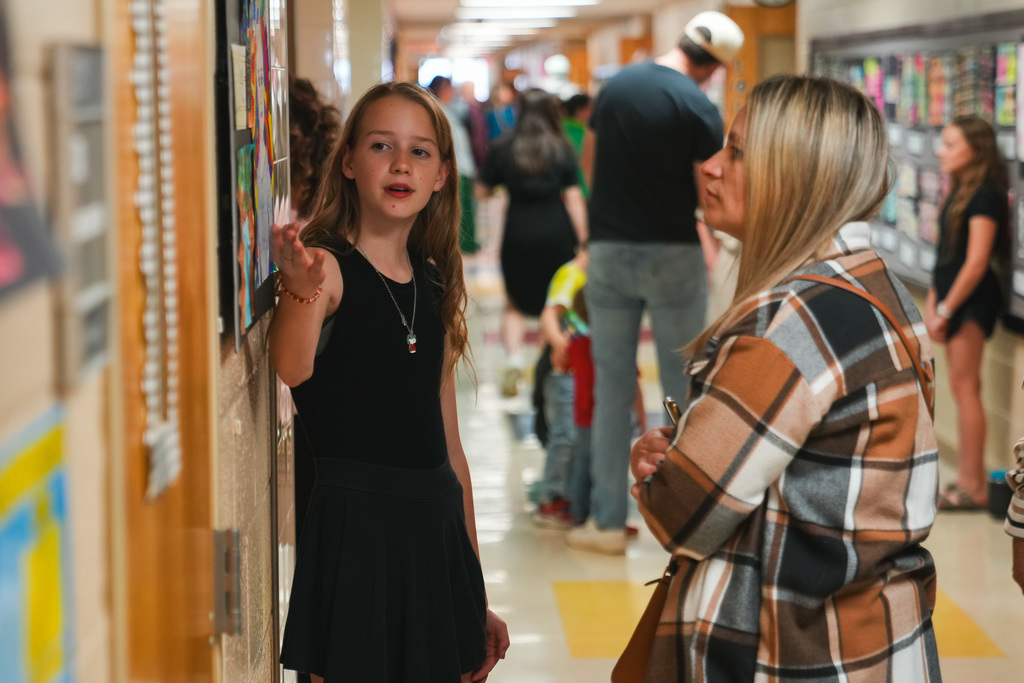 A young girl points at art and talks to an adult woman in a school hallway with other groups of people doing the same
