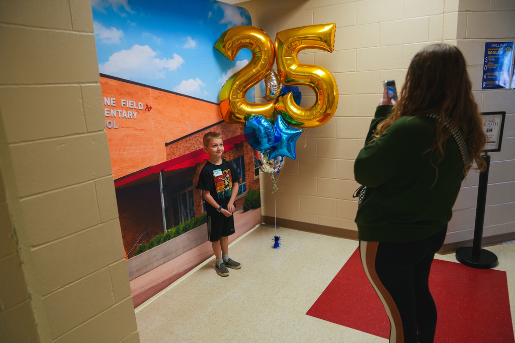 A young boy posses for a picture from an adult woman in front of a picture of Eugene Field A+ Elementary with a balloon in the shape of a 25