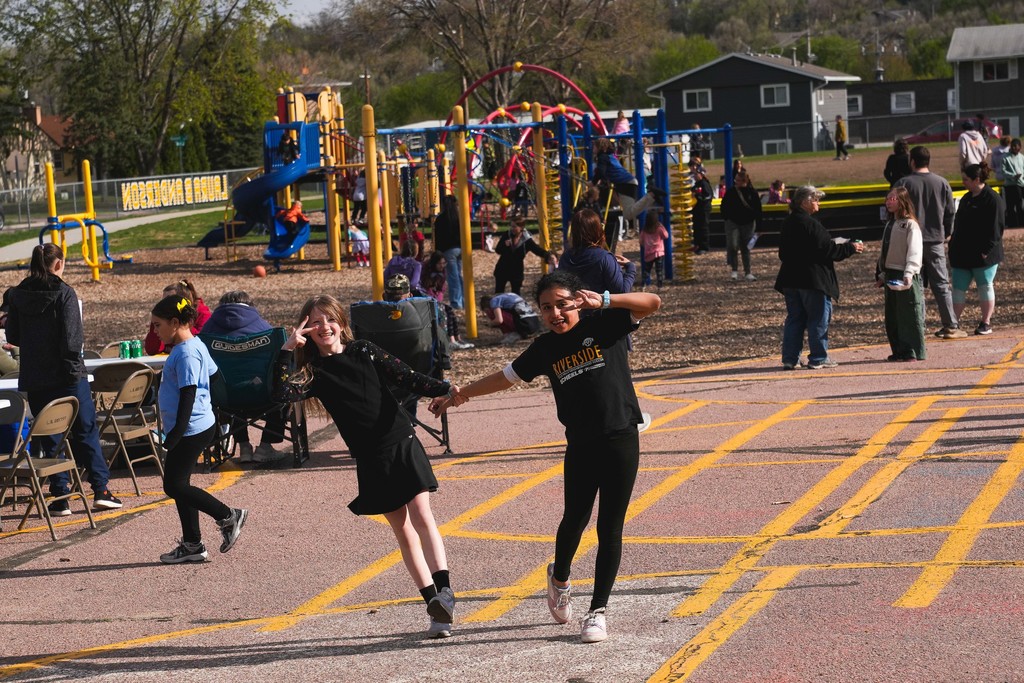 Two girls holding hand show a peace sign to the camera as kids play on a playground in the background
