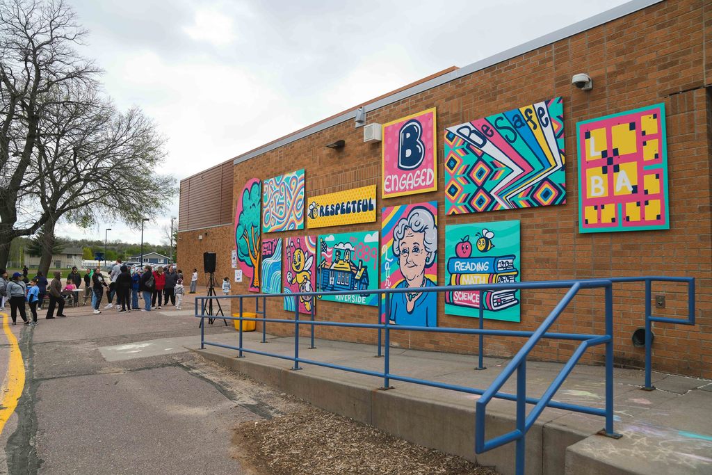 Panels of a colorful painted mural on the side of a school as a crowd gathers in the background