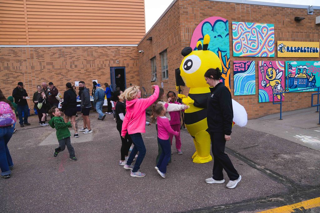 A bee mascot high fives young children on the blacktop of a school playground