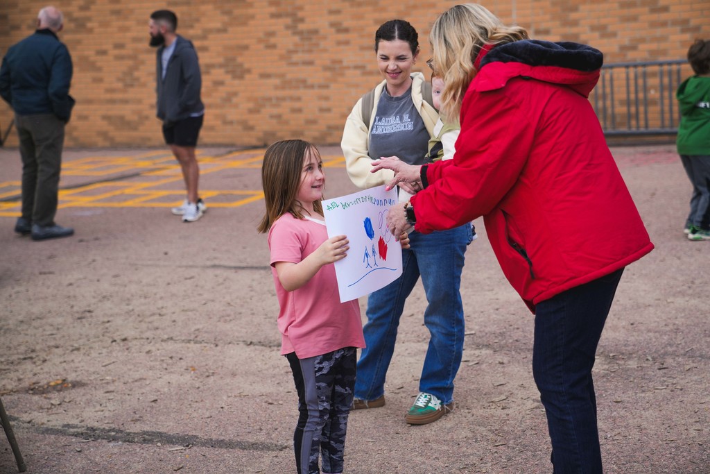 A little girl gives a hand drawn pictures to an adult woman as a mom holding a baby look on in the background