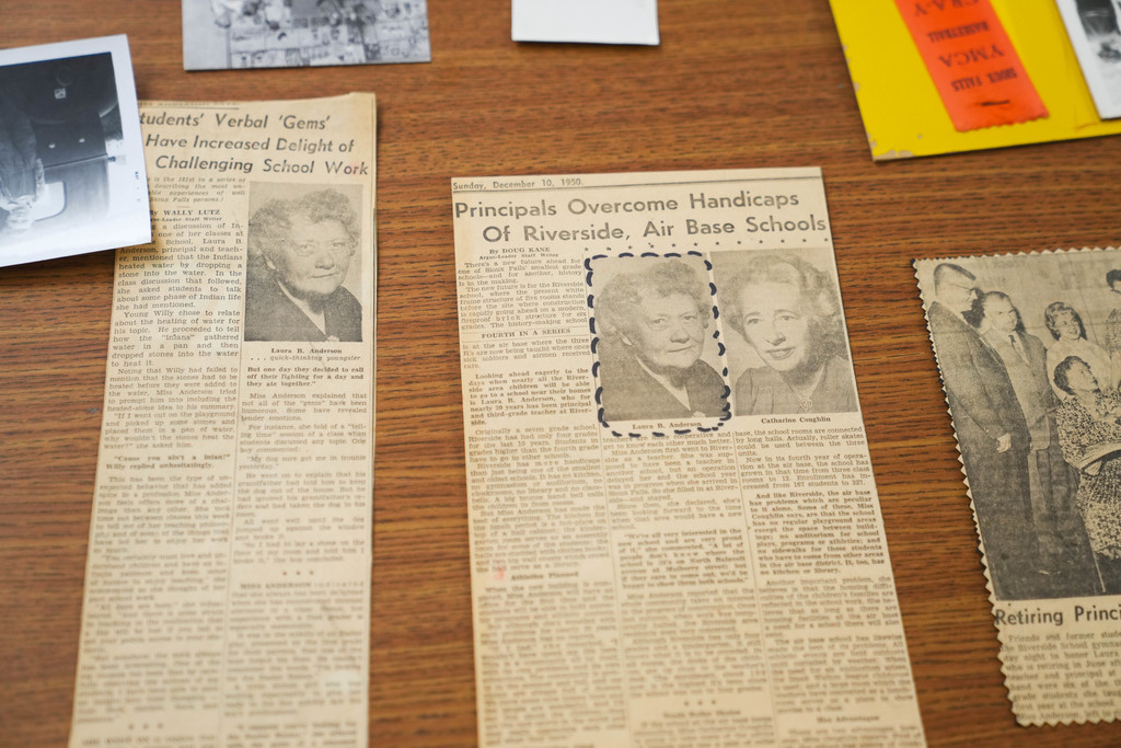 An old newspaper clipping lies on a table. The clipping says "Principles Overcome Handicaps of Riverside, Air Base Schools"