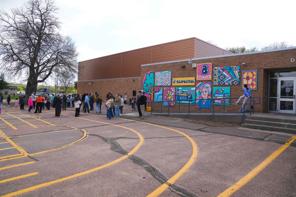 A wide photo of a crowd on a school blacktop with a colorful panel mural on the side of a school